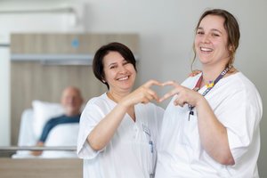 2 Pflegerinnen formen gemeinsame ein Herz mit ihren Händen, hinter ihnen ein Patient im Krankenhausbett