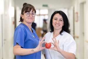 2 Pflegerinnen halten ein rotes Plastik Herz gemeinsam hoch und lächeln in die Kamera