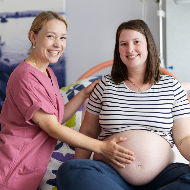 Eine schwangere Frau sitzt auf einem Bett, während eine Hebamme lächelnd ihren Bauch berührt und betreut.