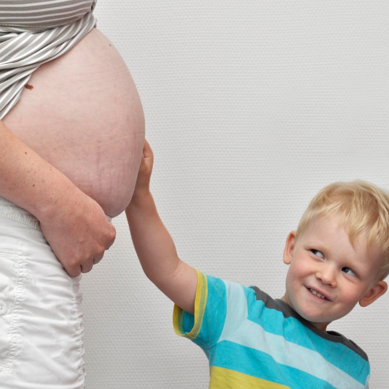 Kleines Kind legt die Hand auf den Bauch einer schwangeren Frau und schaut dabei lächelnd nach oben.