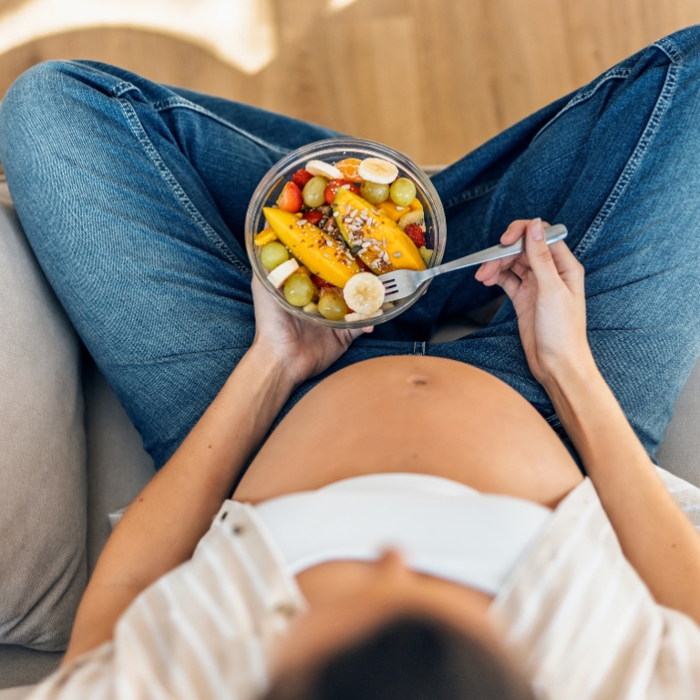 Schwangere Frau sitzt entspannt und isst aus einer Schale mit frischem Obst.
