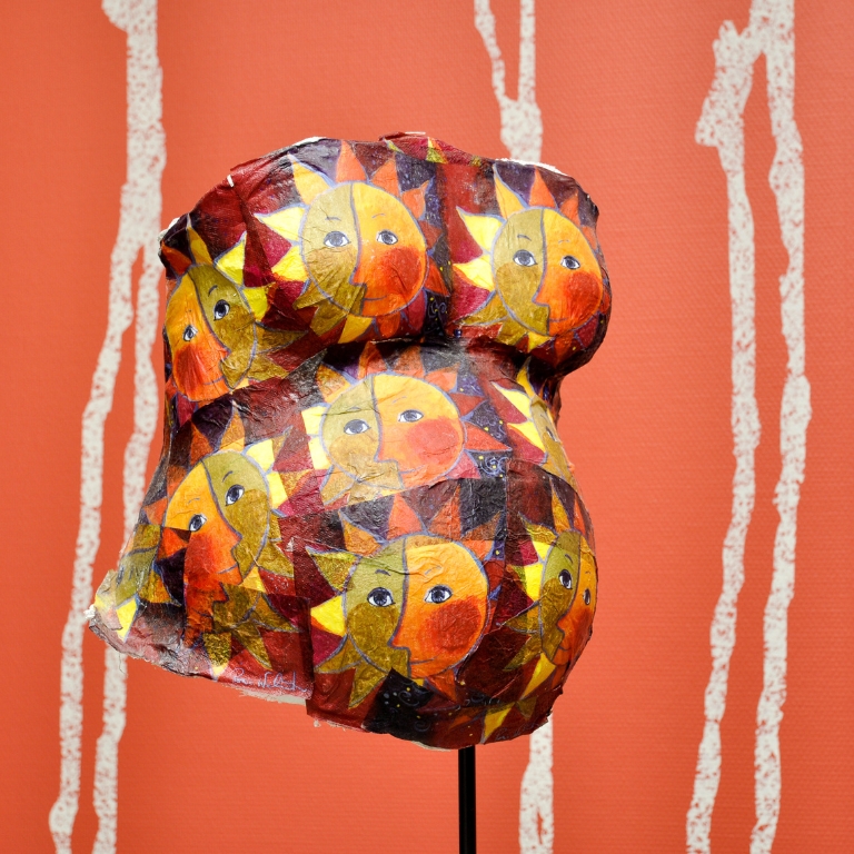 Bemalter Gipsabdruck eines Schwangerschaftsbauchs mit buntem Sonnenmuster auf einem Ständer vor orangem Hintergrund.