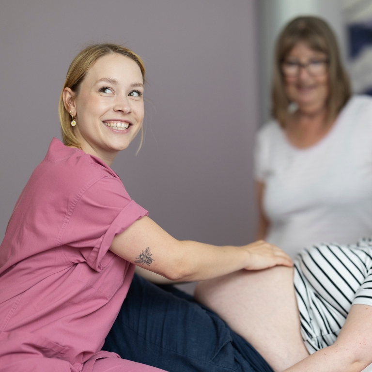 Hebamme in pinker Kleidung sitzt neben einer schwangeren Frau und berührt ihren Bauch während einer Beratungssituation.