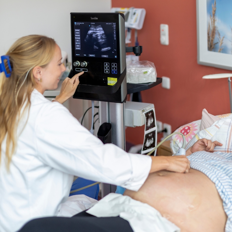 Medizinische Fachkraft führt eine Untersuchung am Bauch einer schwangeren Frau durch, während ein Ultraschallgerät im Hintergrund zu sehen ist.