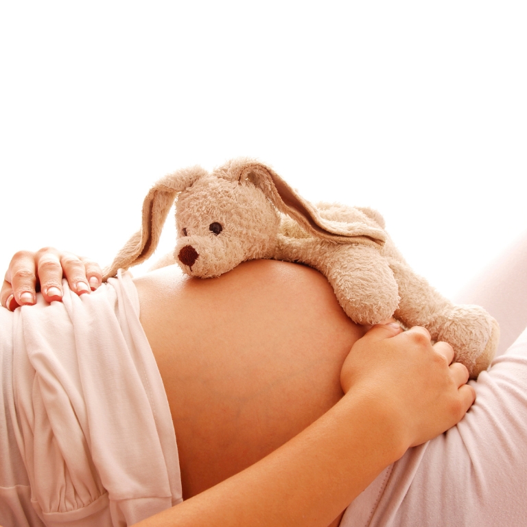 Schwangere Frau hält ihren Bauch, auf dem ein kleines Stofftier liegt.