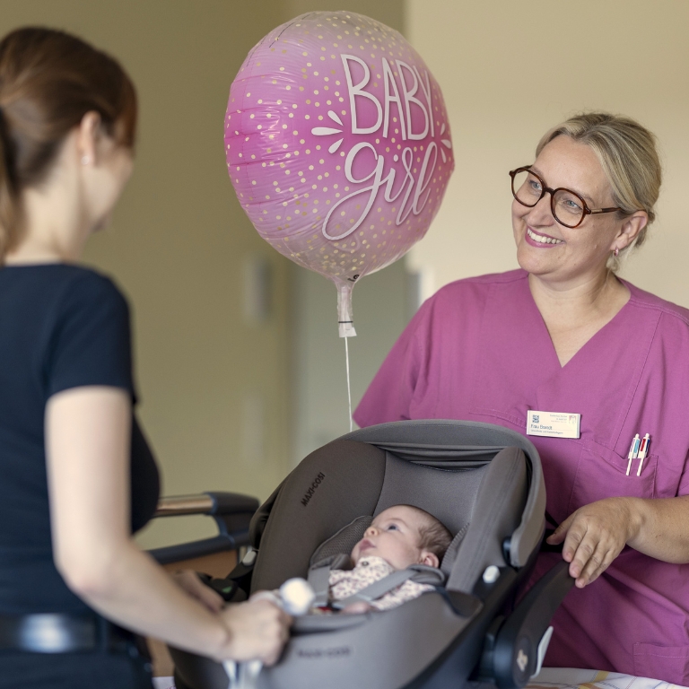 Eine Frau im medizinischen Outfit spricht mit einer Mutter, während ein Baby in einer Babyschale liegt.