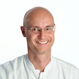 Arzt in weißer Arbeitskleidung lächelt vor neutralem Hintergrund.