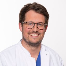 Arzt in weißer Arbeitskleidung lächelt vor neutralem Hintergrund.