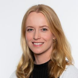 Porträt einer lächelnden Frau mit blondem, schulterlangem Haar vor hellem Hintergrund.