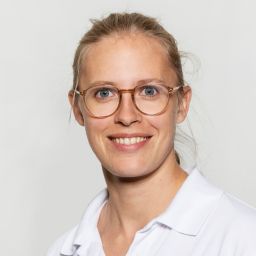 Ärztin in weißer Arbeitskleidung lächelt vor neutralem Hintergrund.