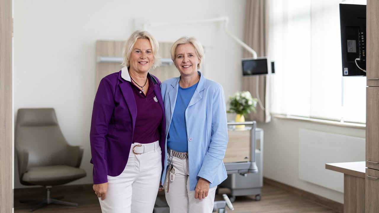 2 schick-gekleidete Mitarbeiterinnen stehen Arm-in-Arm in einem modernen Patientenzimmer und lächeln in die Kamera
