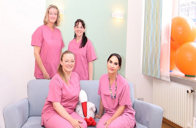 4 Hebammen und Pflegerinnen posieren an einem Sofa und lächeln einladend in die Kamera