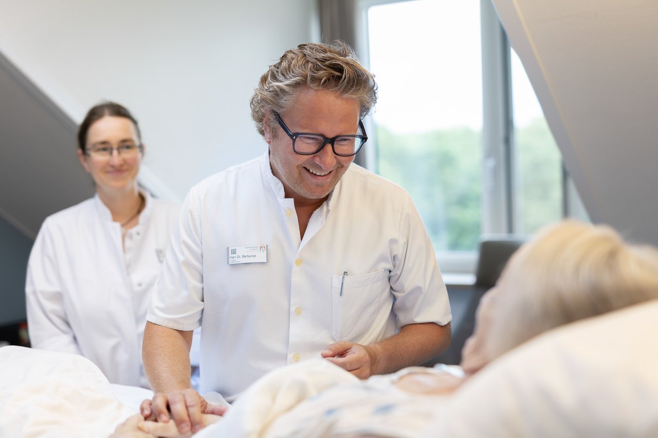 Ein Arzt spricht mit einer älteren Dame, die im Patientenbett liegt, im Hintergrund eine weitere Ärztin