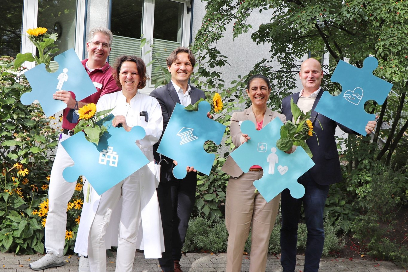Fünf Personen stehen im Freien und halten symbolische Puzzleteile eines gemeinsamen Projekts in den Händen.