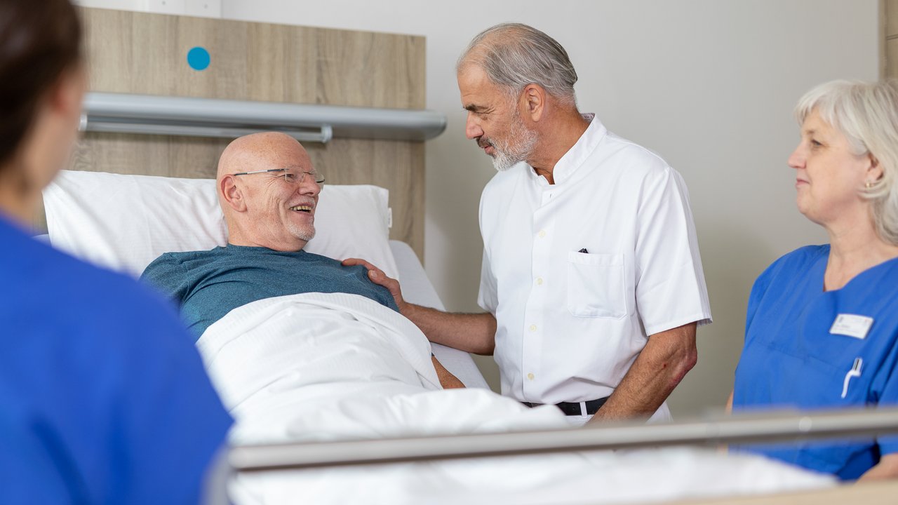 Ein Arzt spricht eingehend mit einem Patienten, der im Krankenhausbett liegt. Herum stehen 2 Pflegerinnen