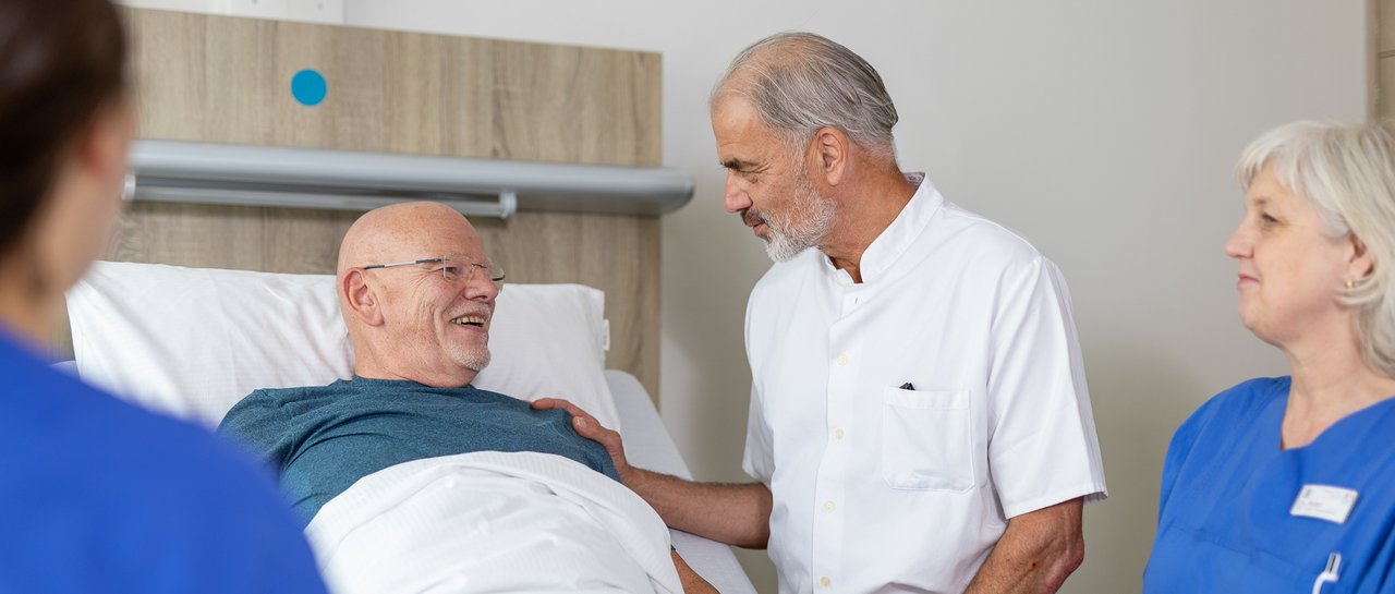Ein Arzt spricht eingehend mit einem Patienten, der im Krankenhausbett liegt. Herum stehen 2 Pflegerinnen