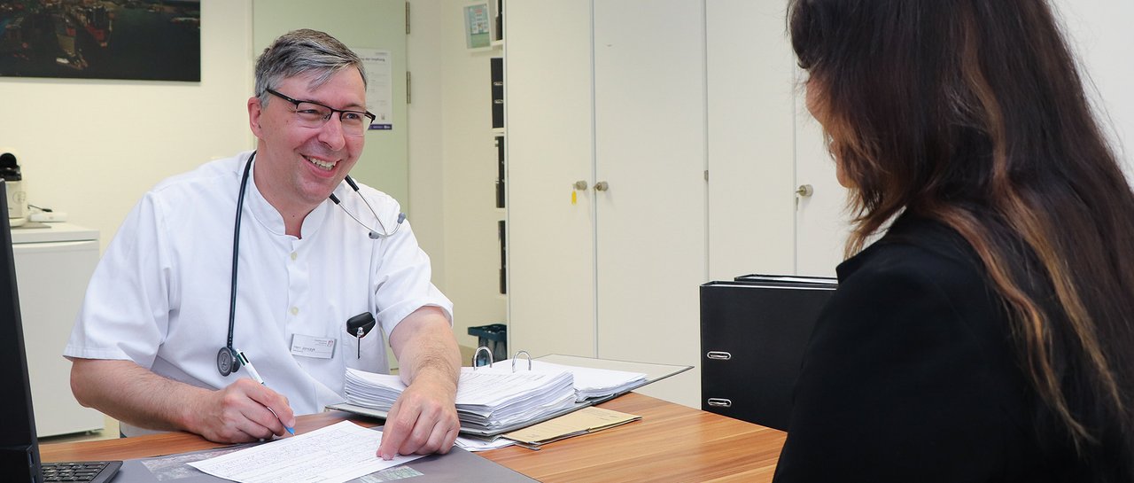Ein Arzt sitzt am Schreibtisch und erklärt einer Patientin etwas lächelnd