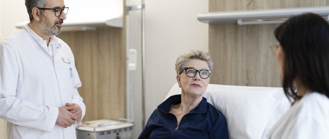 Eine Pflegerin und ein Arzt stehen am Patientenbett einer älteren Dame und sprechen mit ihr.