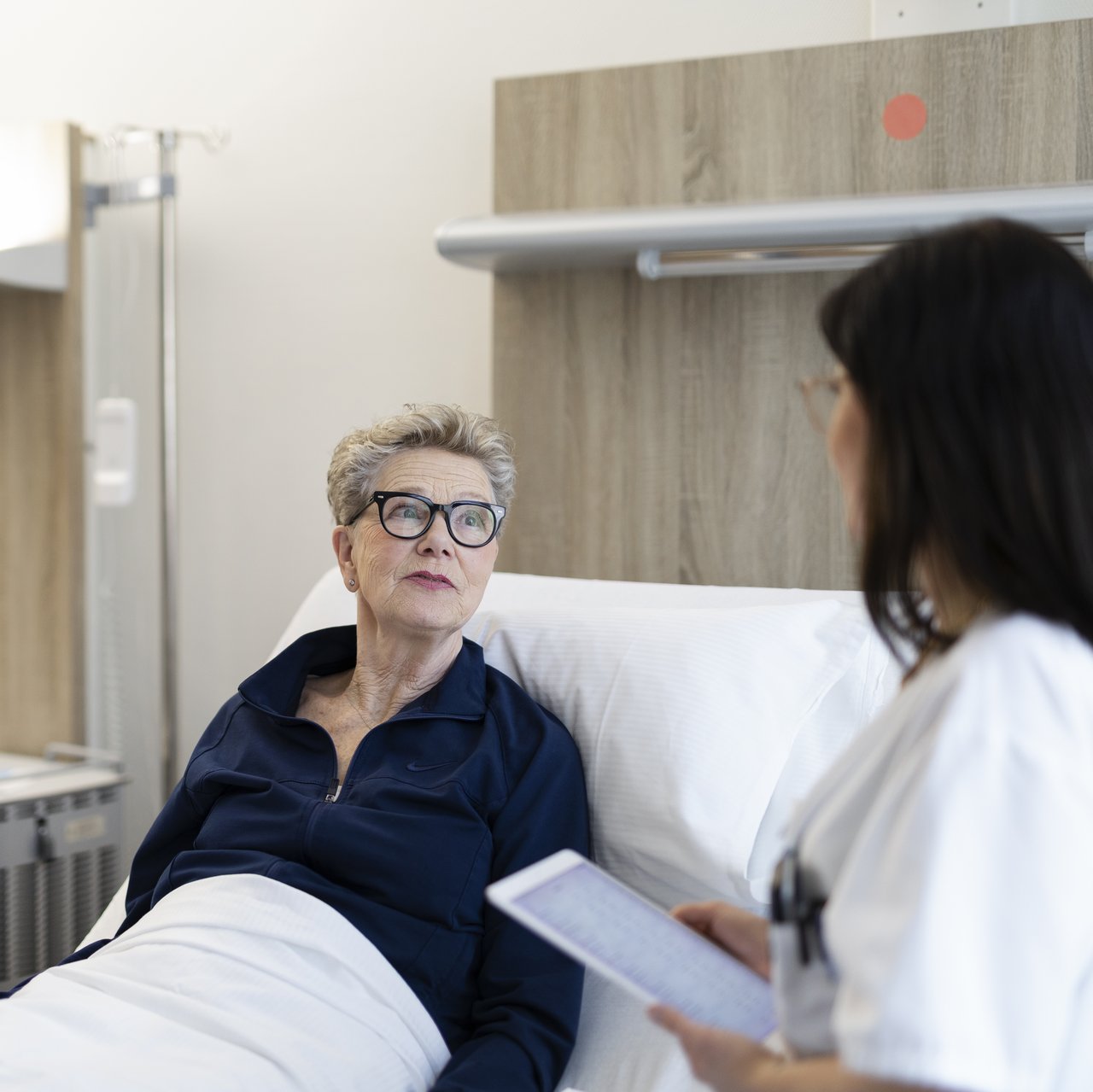 Ein Oberarzt spricht mit einer Pflegerin und einer älteren Patientin, die im Krankenhausbett liegt.
