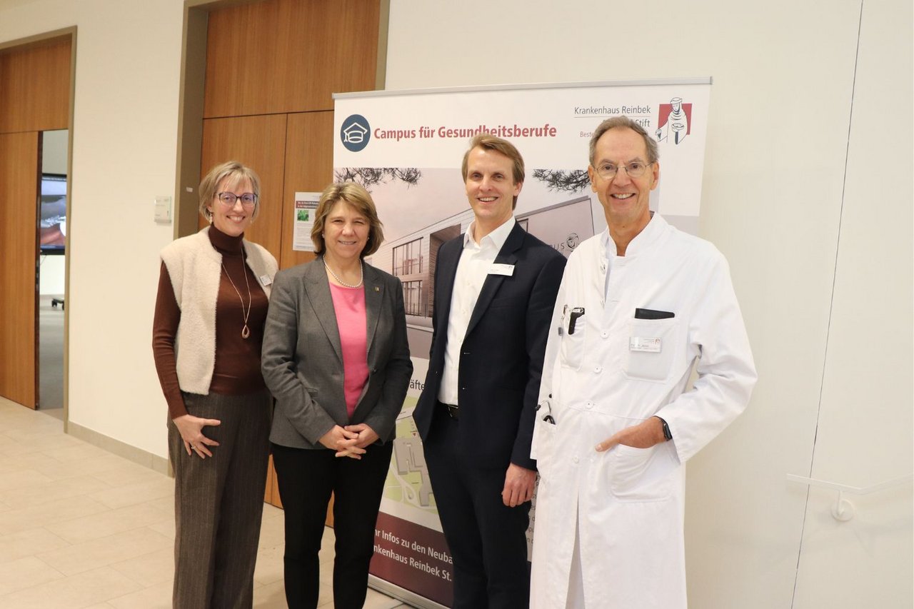 Vier Personen stehen lächelnd im Eingangsbereich eines Gebäudes vor einem  großem Plakat des „Campus für Gesundheitsberufe“.
