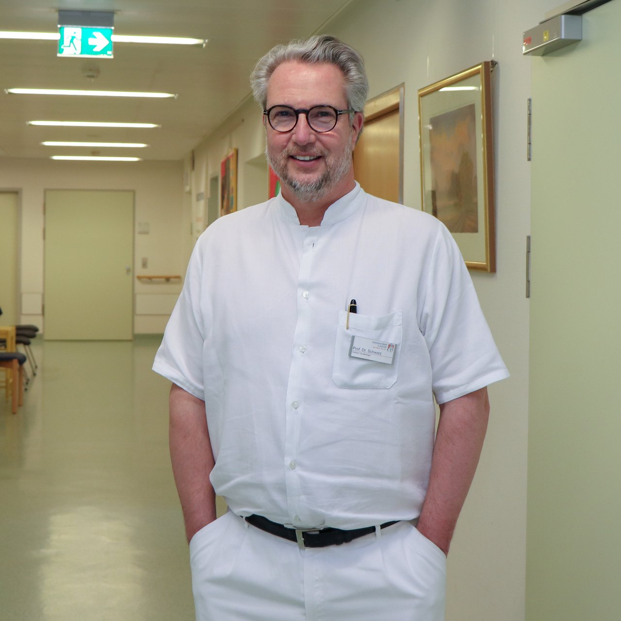 Ein Chefarzt auf dem Krankenhausflur. Er hat die Hände in der Hosentasche und lächelt in die Kamera