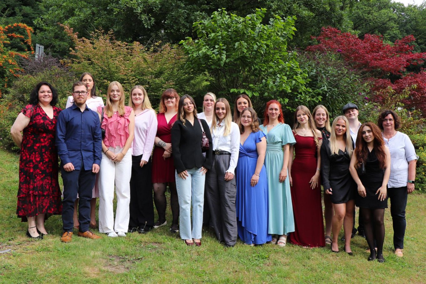 16 Absolventinnen und Absolventen der generalistischen Pflegeausbildung stehen mit zwei Begleitpersonen für ein Gruppenfoto im Garten des Krankenhauses Reinbek. Die Gruppe ist festlich gekleidet und steht auf einer Wiese vor Bäumen und Sträuchern.