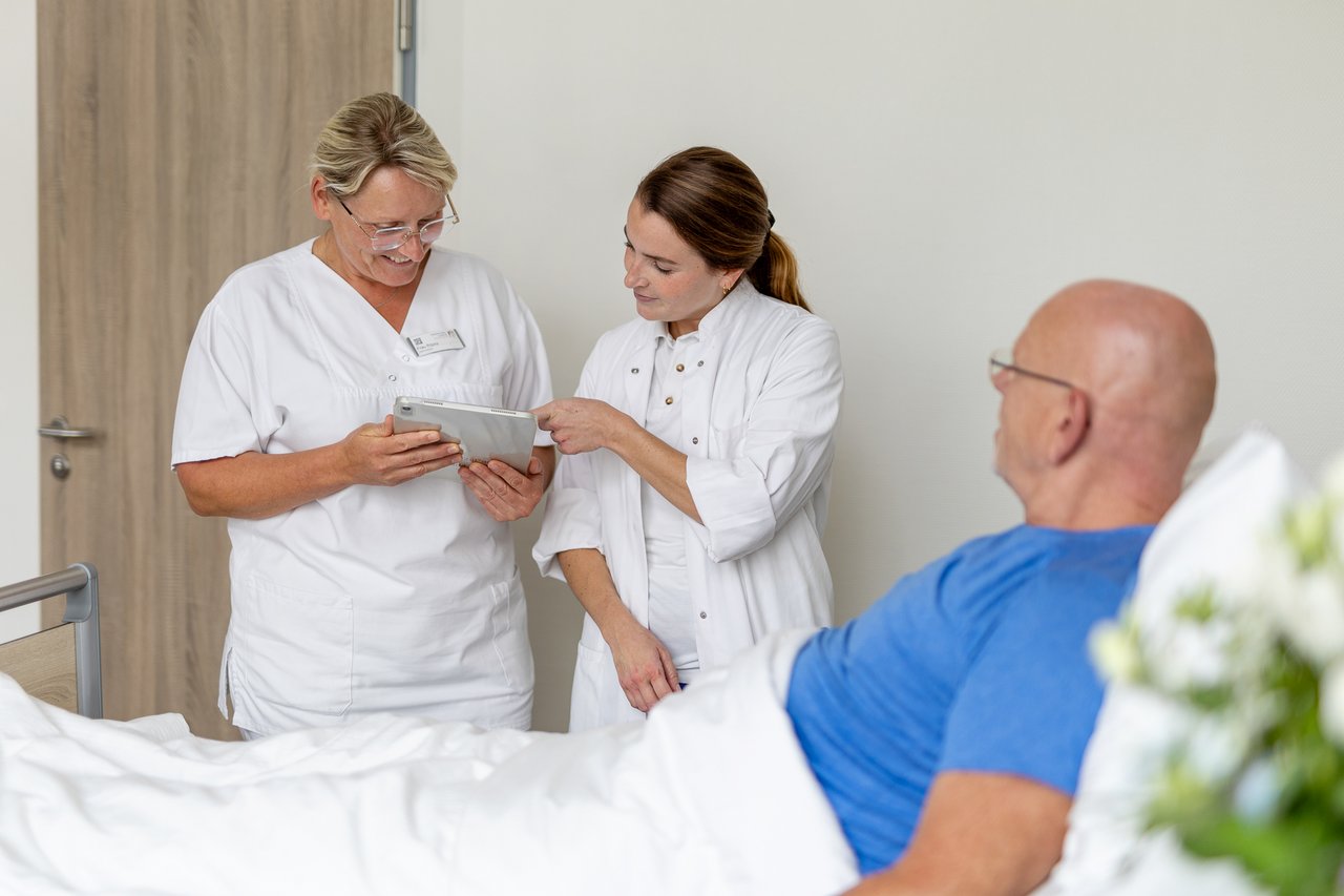 Eine Pflegerin und Ärztin schauen auf ein Tablet, neben ihnen ein Patient im Bett