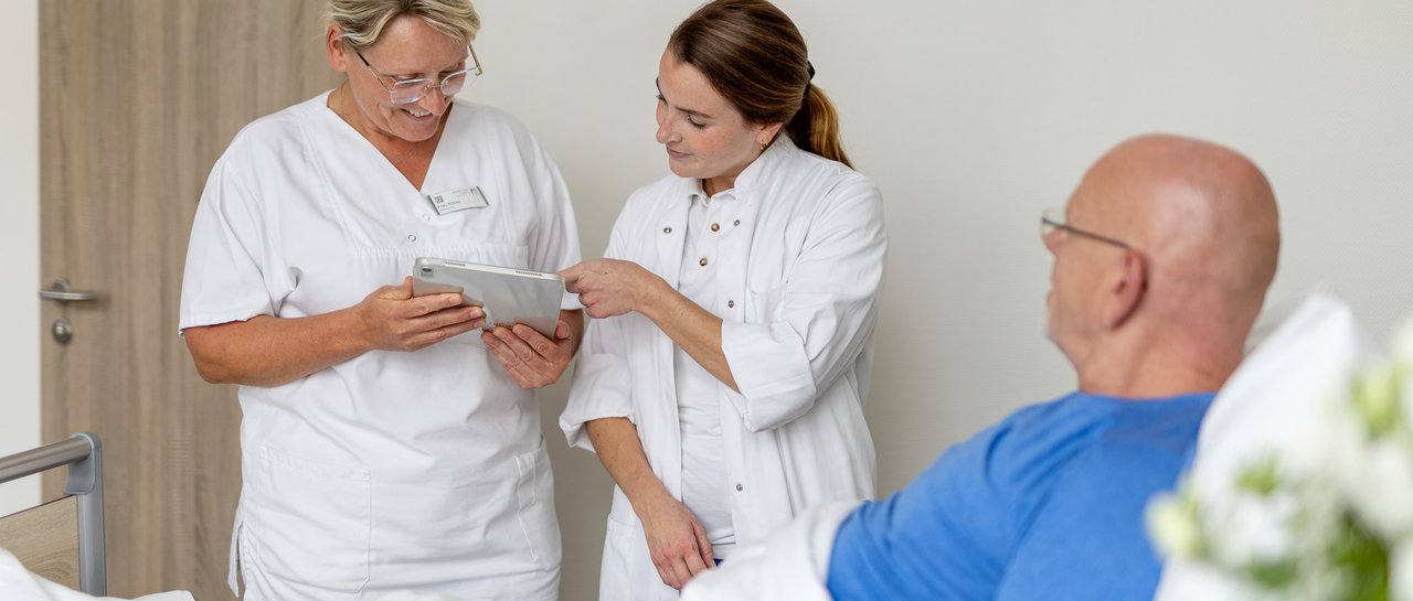 Eine Pflegerin und Ärztin schauen auf ein Tablet, neben ihnen ein Patient im Bett