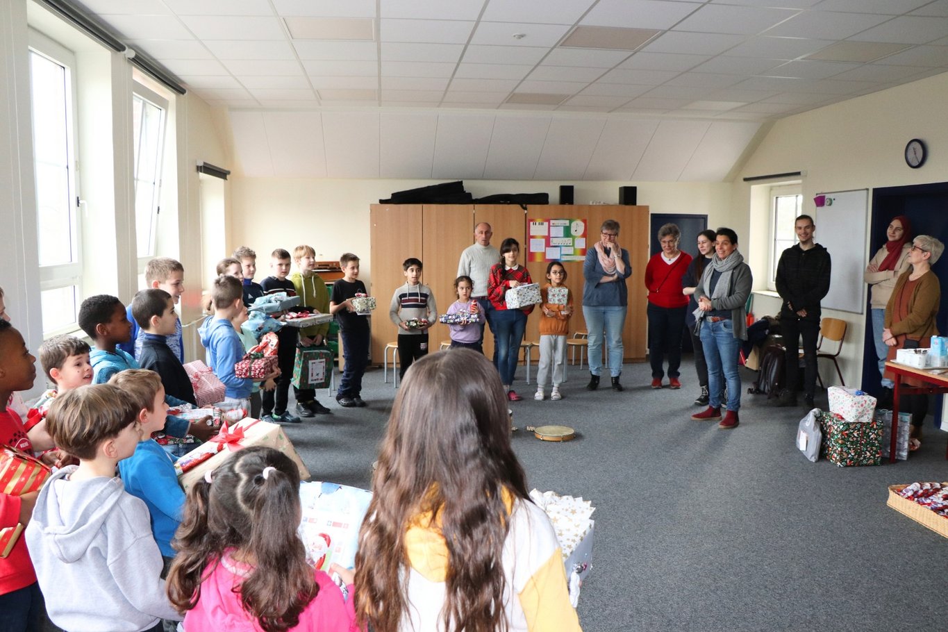 Kinder stehen in einem Klassenraum im Kreis, halten Weihnachtsgeschenke in den Händen und freuen sich gemeinsam, während mehrere Erwachsene am Rand zuschauen.