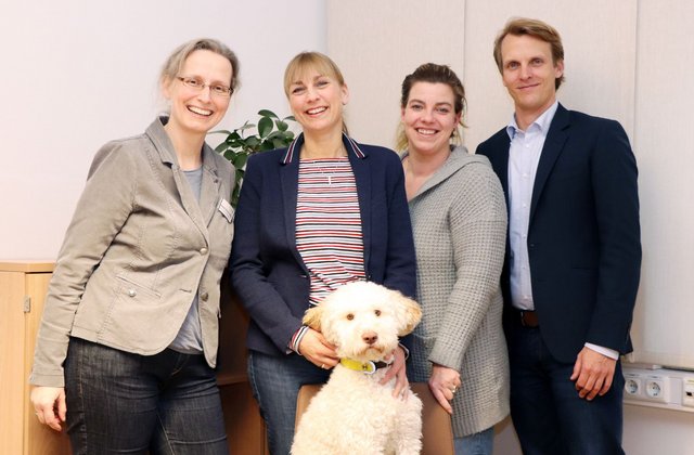 Vier Mitarbeitende des Krankenhauses Reinbek St. Adolf-Stift stehen zusammen mit einem kleinen weißen Hund vor der Kamera und lächeln.