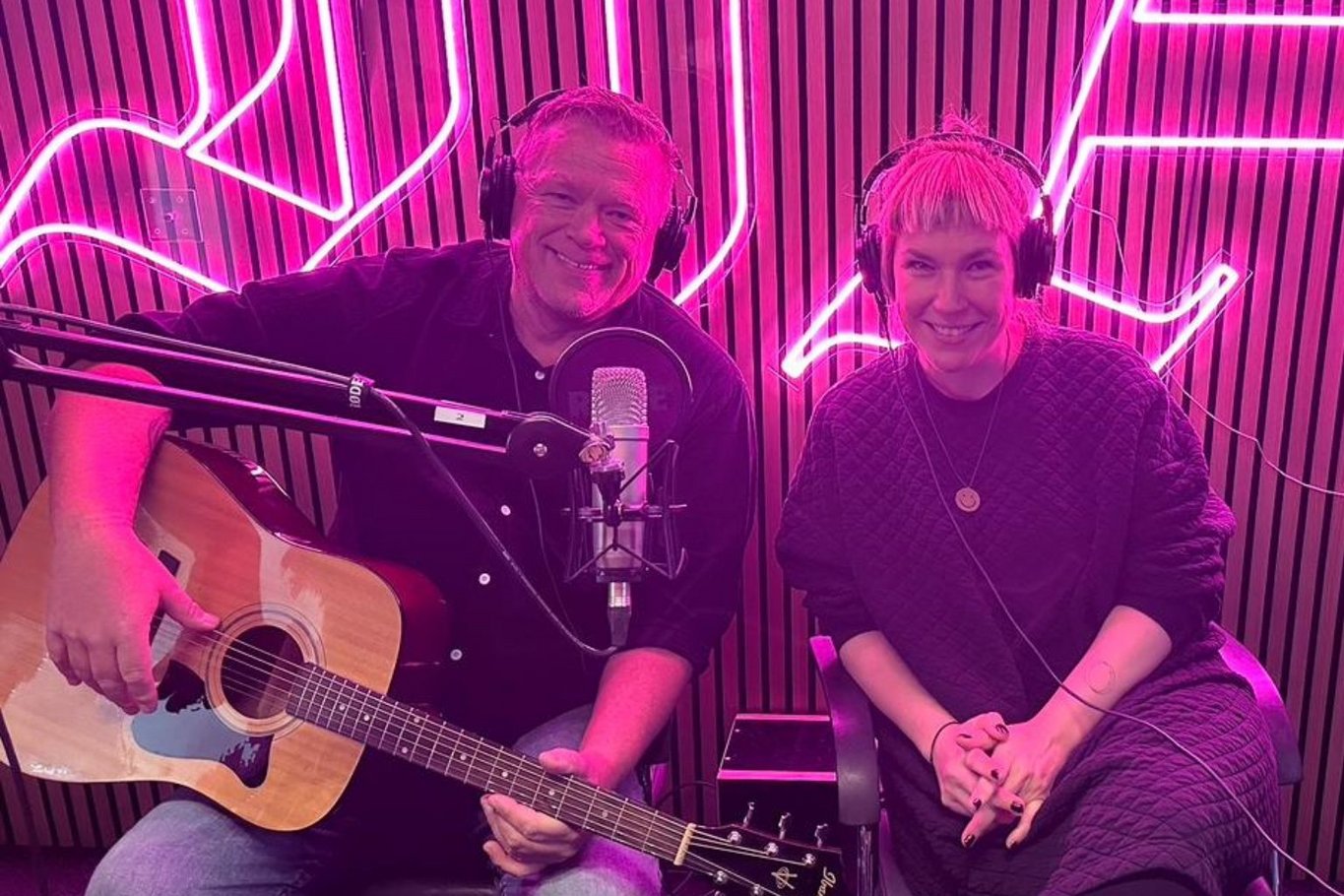 Zwei Personen sitzen mit Kopfhörern in einem Podcaststudio vor einem Mikrofon, eine Person hält eine Gitarre, im Hintergrund leuchtet eine pinke Neonbeleuchtung.