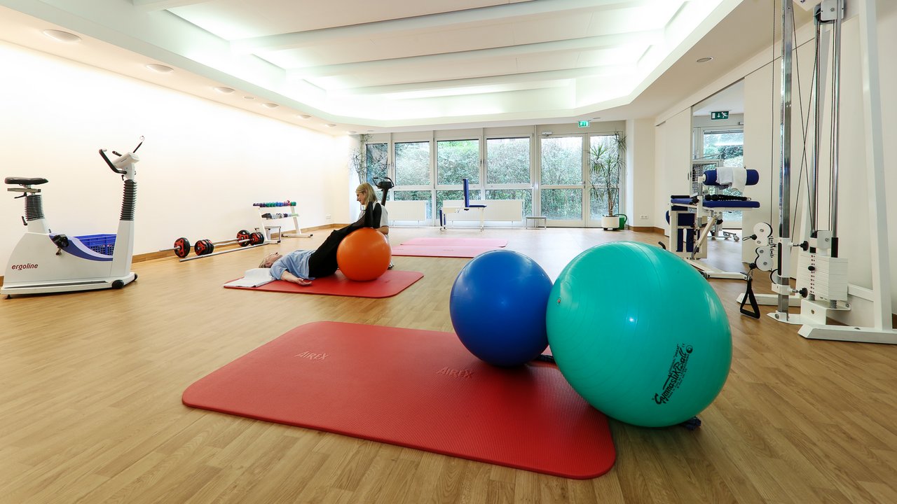 Ein Bild einer Physiotherapie-Raumes mit vielen Gymnastikbällen und einer Patientin die im Hintergrund eine Übung mit dem Gymnastikball macht