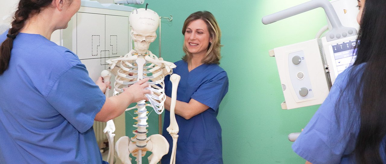 Eine Praxisanleiterin der Radiologie zeigt Azubis mit einem Skelett wie man einen Röntgen macht.