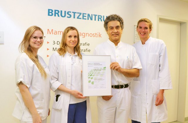Vier Mitarbeitende des Brustzentrums stehen vor einer Wand mit der Aufschrift „Brustzentrum“ und präsentieren ein Zertifikat der Deutschen Krebsgesellschaft.