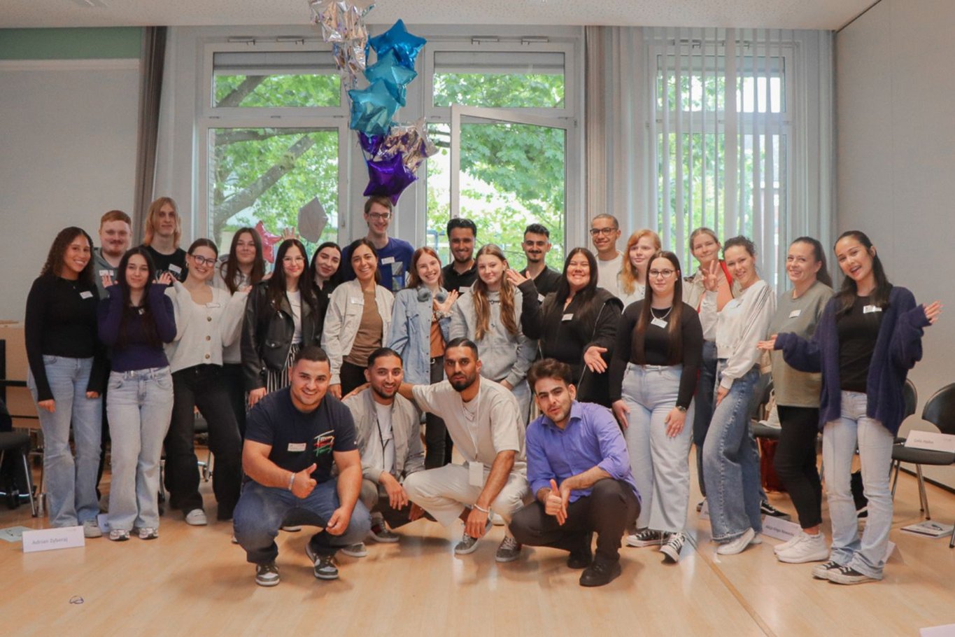 Gruppenfoto von etwa 26 jungen Erwachsenen in einem hellen Seminarraum vor großen Fenstern. Einige stehen, vier Männer knien vorne. Mehrere Personen tragen Namensschilder und winken in die Kamera; im Hintergrund hängen bunte Sternballons.