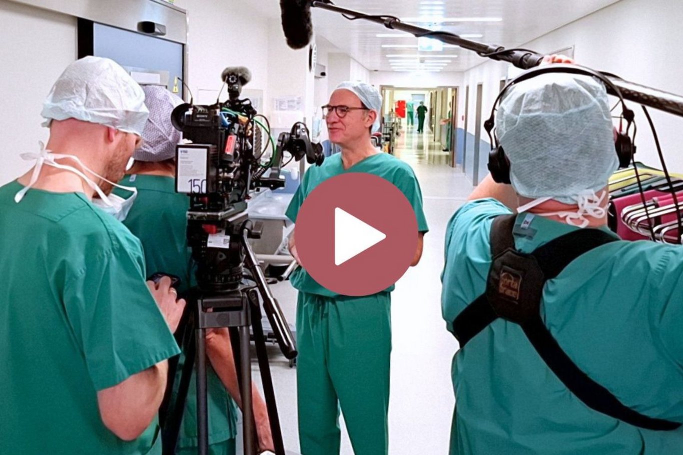 Ein Arzt in OP-Kleidung wird in einem Krankenhausflur von einem Fernsehteam interviewt; Kamera und Mikrofon sind auf ihn gerichtet, während im Hintergrund Krankenhauspersonal durch den Flur geht.