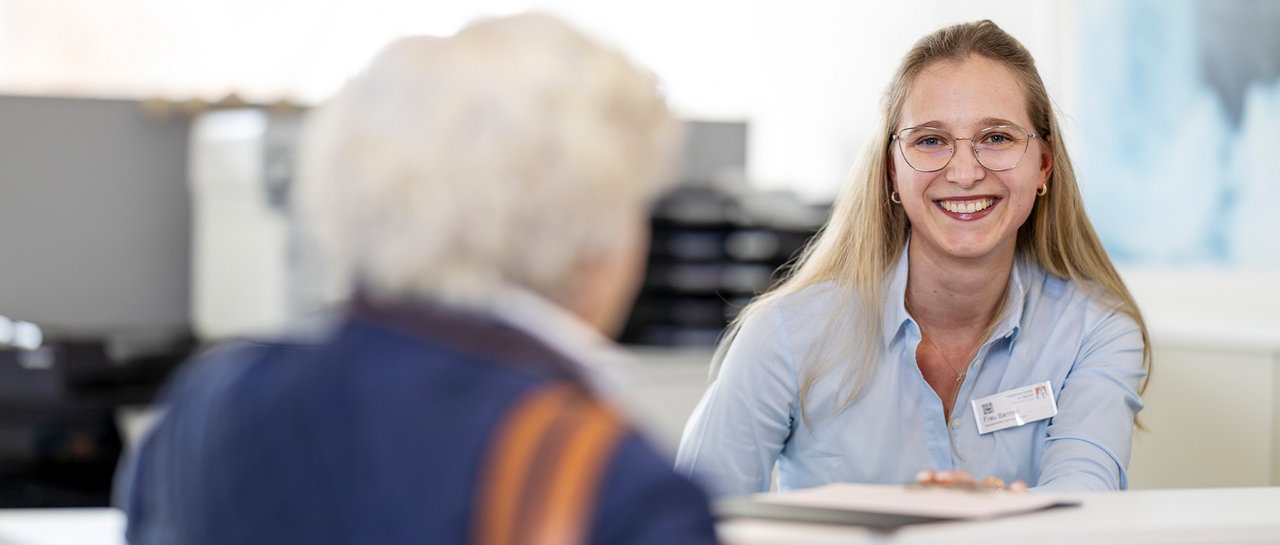 Eine junge Dame sitzt am Empfang, bedient eine ältere Dame und lächelt freundlich in die Kamera
