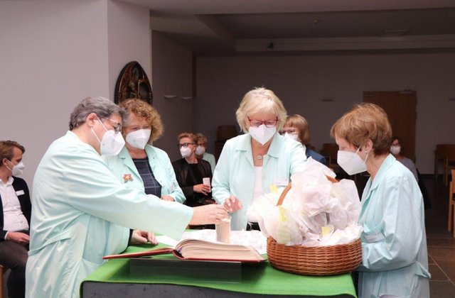 Mehrere Frauen der „Grünen Damen“ stehen während einer Andacht in einem Krankenhausraum an einem Tisch mit Kerze, aufgeschlagenem Buch und einem Korb mit Tüchern und führen gemeinsam ein Ritual durch.