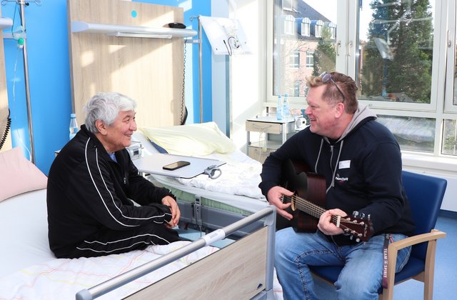 Ein Patient sitzt auf seinem Krankenhausbett und hört einem Mann zu, der ihm in einem hellen Patientenzimmer auf einer Gitarre vorspielt. Beide lächeln sich an.