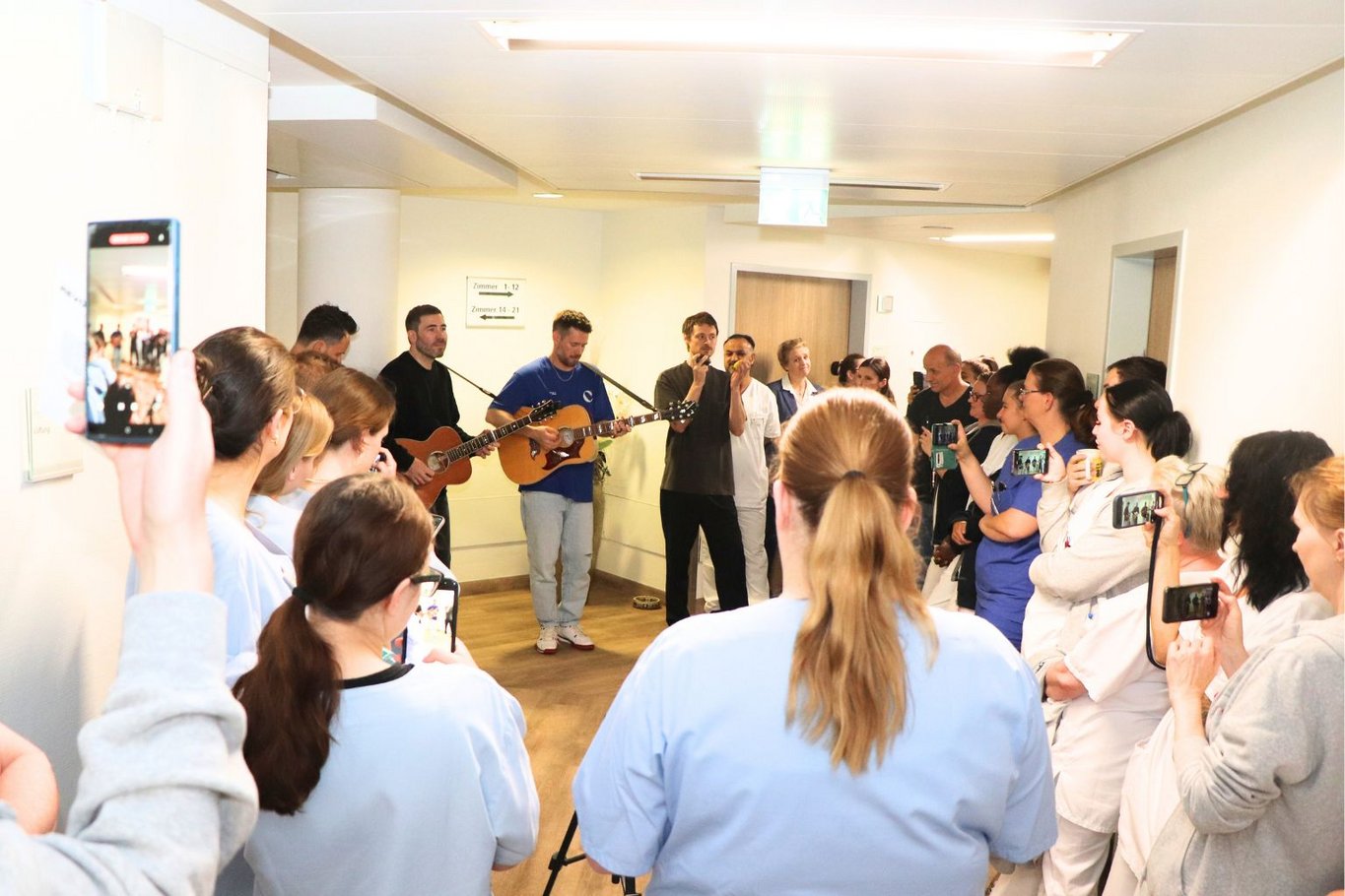 Mitarbeitende des St. Adolf-Stiftes stehen auf einem Krankenhausflur und hören einem Konzert der Band Revolverheld zu. Musiker spielen Gitarre und singen, während Pflegekräfte und andere Mitarbeitende in Arbeitskleidung zuhören und filmen.