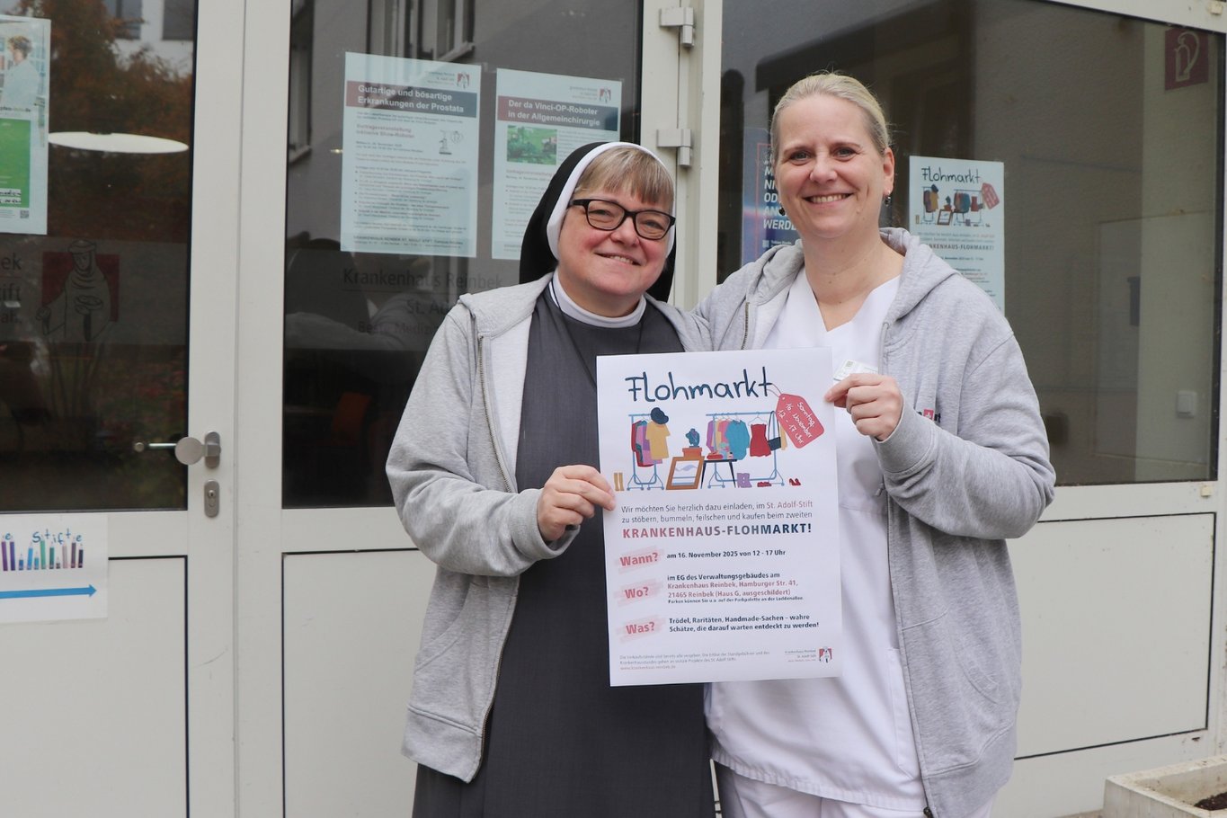 Vor dem Eingang der Aula stehen Schwester Luise und Yvonne Hillebrand vom Organisationsteam und halten ein Plakat hoch, das den Flohmarkt am 16. November im St. Adolf-Stift ankündigt.