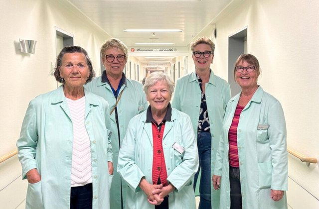 Fünf Frauen in hellgrünen Kitteln stehen gemeinsam auf einem Flur im Krankenhaus. Sie blicken mit einem Lächeln in die Kamera.