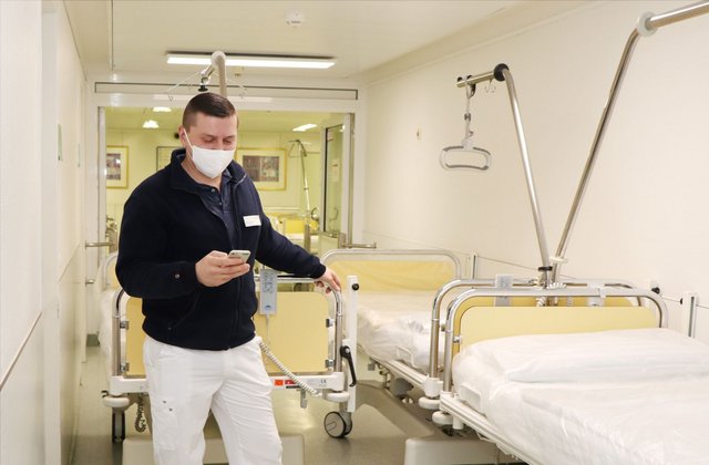 Ein Krankenhausmitarbeiter mit Mundschutz steht im Flur neben einem Patientenbett und schaut auf ein mobiles Gerät, über das er einen neuen Transportauftrag erhält.