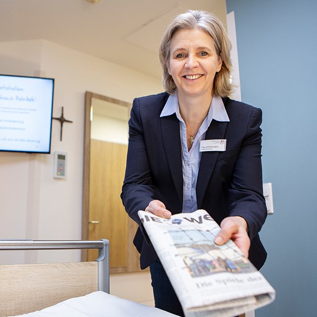 Eine Mitarbeiterin übergibt dem Patienten hinter der Kamera im Krankenhausbett eine Broschüre
