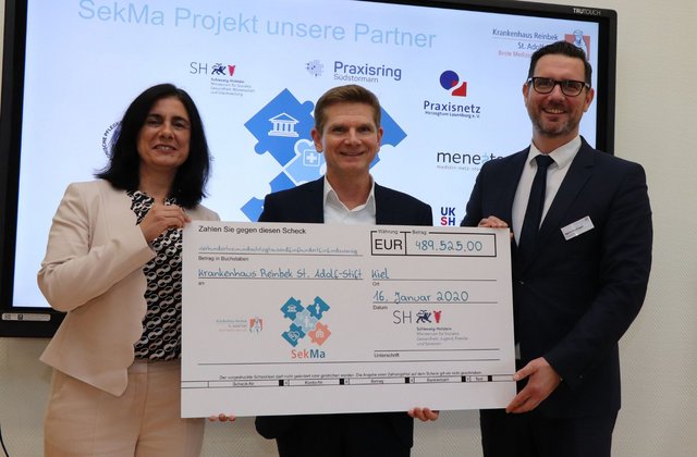 Drei Personen stehen vor einer Präsentationswand und halten gemeinsam einen großen symbolischen Scheck über knapp 490.000 Euro für das Krankenhaus Reinbek St. Adolf-Stift in die Kamera.
