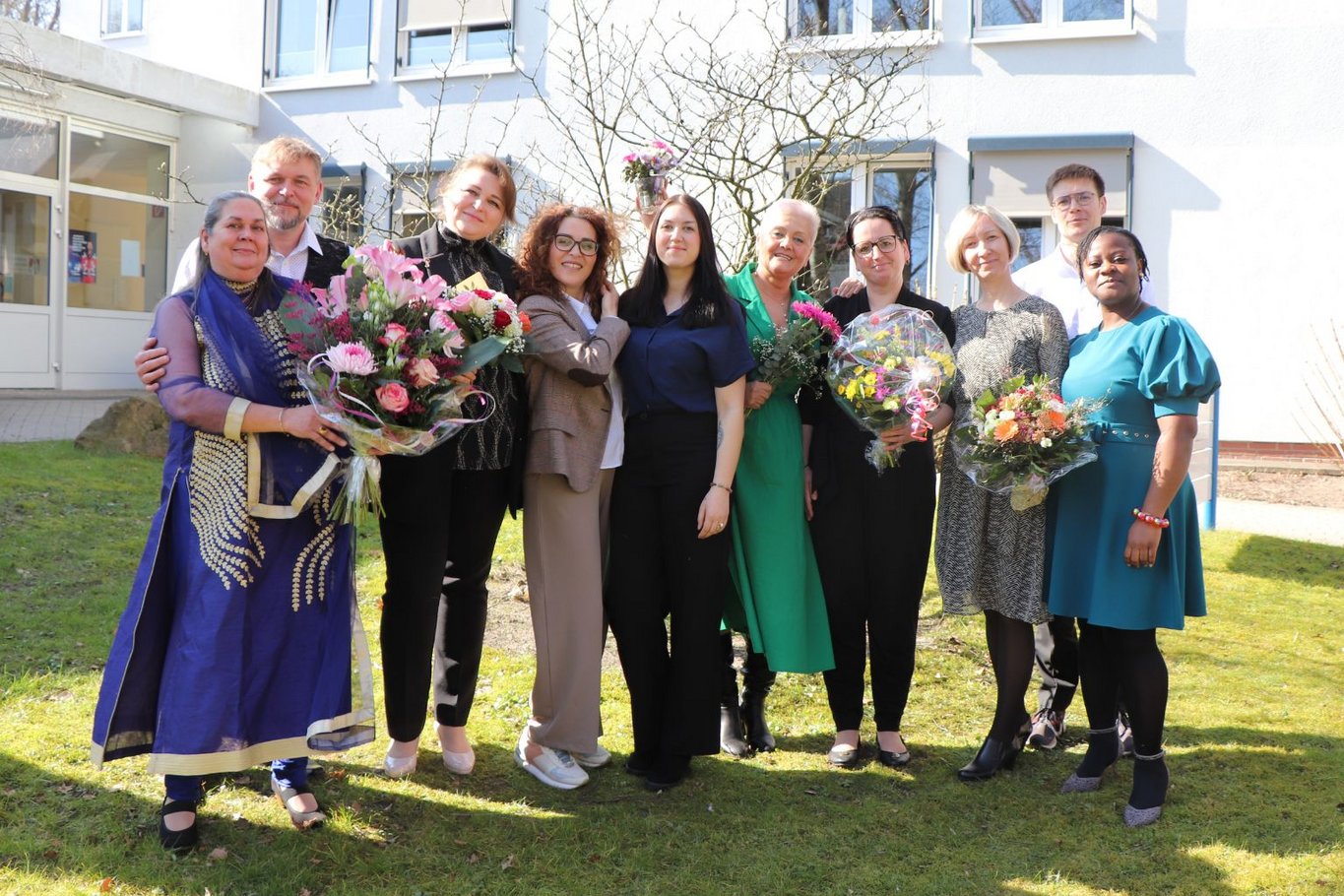 Gruppe von Absolventinnen und Absolventen eines Pflegekurses steht im Freien vor einem Gebäude und hält Blumensträuße nach bestandener Prüfung.