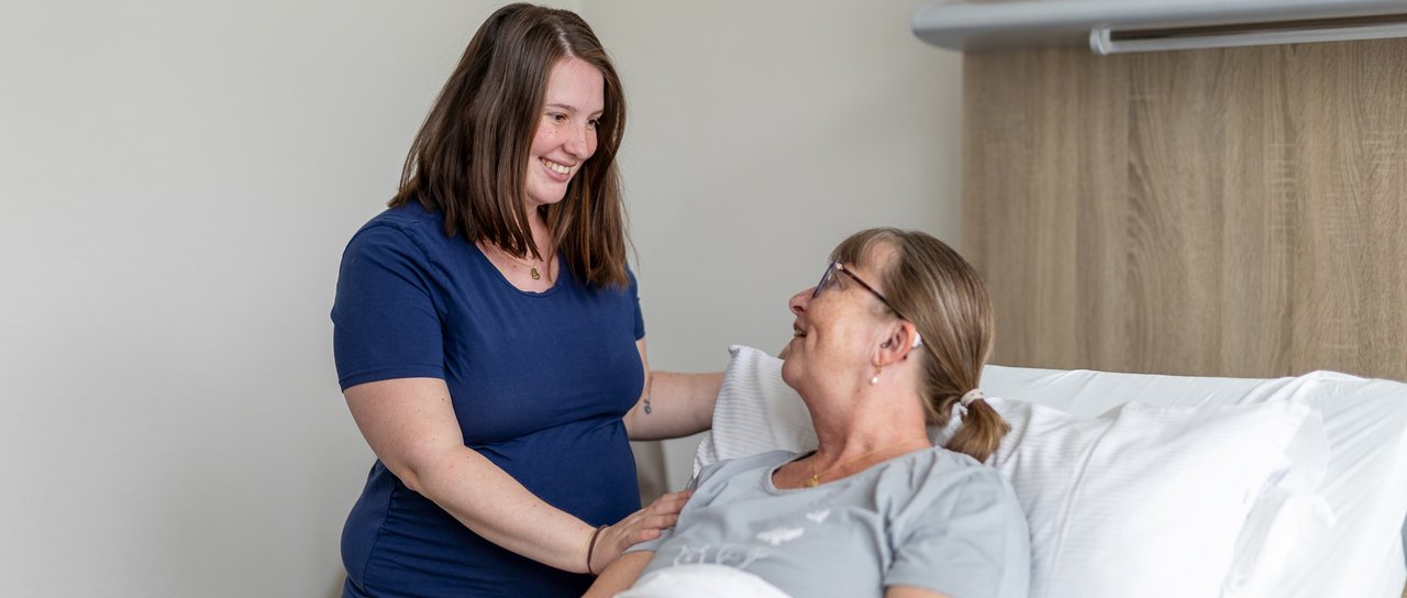 Eine Tochter steht am Patientenbett ihrer Mutter und lächelt sie an