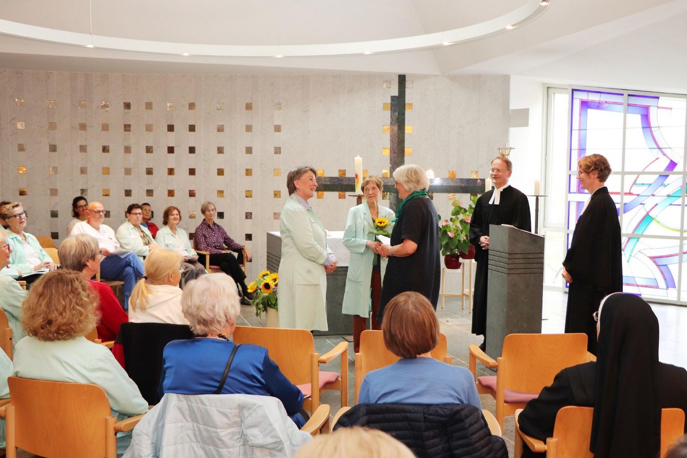 Veranstaltung in der Kapelle des Krankenhauses Reinbek: Mehrere Frauen stehen vor dem Altar und sprechen miteinander, während Gäste auf Stühlen sitzen und zuschauen.