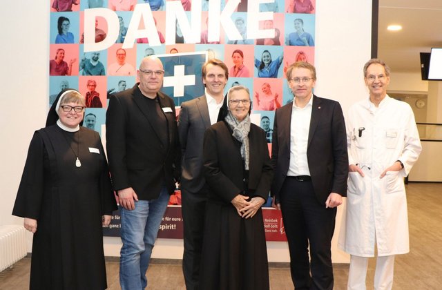 Sechs Personen aus Gesundheitsminister, Leitung, Pflege und Medizin des St. Adolf-Stifts stehen gemeinsam vor einem ‚Danke‘-Plakat mit Porträts von Mitarbeitenden.