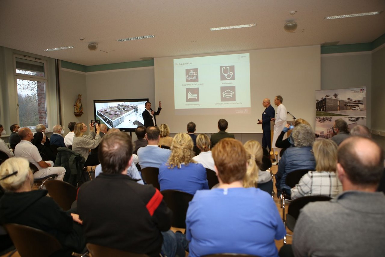 Mehrere Personen sitzen in einem Veranstaltungsraum und verfolgen eine Präsentation, während vorne Referenten über Fördermaßnahmen im Gesundheitswesen sprechen.